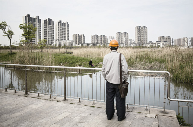 중국 상하이 자딩 지구에서 한 노동자가 연못 건너편에 건설 중인 대규모 아파트 단지를 바라보고 있다. 지난 20여년 동안 중국 대도시 근교 농지 가격이 급등했지만, 중국 정부는 농지의 소유권을 인정하지 않아 농민들은 시장가치로 농지를 매각할 수 없는 상태다. <사진 : 블룸버그>