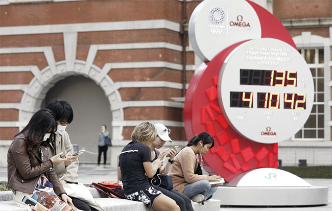 마스크를 쓴 사람들이 3월 11일 수요일 일본 도쿄역 외곽에 있는 도쿄 2020 올림픽의 카운트다운 시계 앞에 앉아 스마트폰을 사용하고 있다. 사진 블룸버그